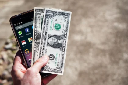 App vay 0 đồng đầu tiên: Close-up of a hand holding US dollar bills and a smartphone outdoors, showcasing financial technology.